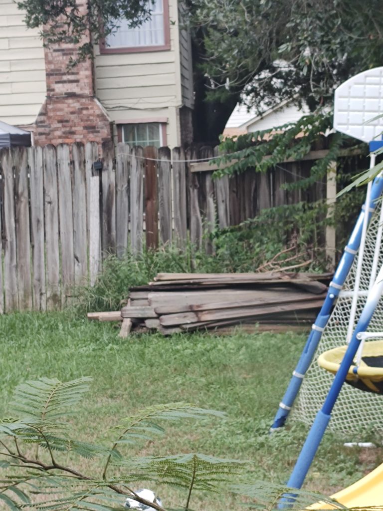 Home 5 Backyard view showing weathered wooden fence, stacked lumber boards on grass, and partial playground equipment